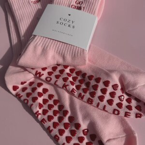 May include: A pair of light pink crew socks featuring the embroidered text "YOU GO GIRL!" in red. The soles are covered in red heart-shaped silicone grips and the text "WOLKENBILDER". A white paper band labeled "COZY SOCKS" is wrapped around the socks.