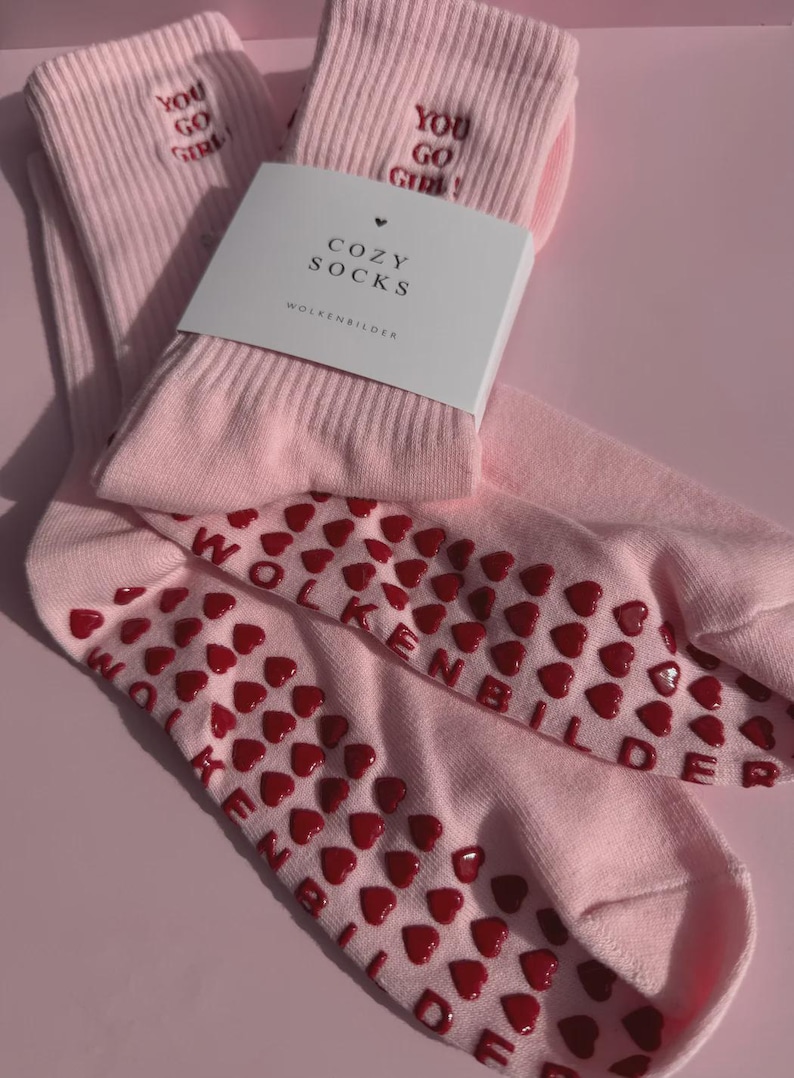 May include: A pair of light pink crew socks featuring the embroidered text "YOU GO GIRL!" in red. The soles are covered in red heart-shaped silicone grips and the text "WOLKENBILDER". A white paper band labeled "COZY SOCKS" is wrapped around the socks.