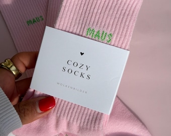 May include: A pair of light pink ribbed socks featuring the green embroidered text "MAUS" on the cuff. The socks are wrapped in a white paper band printed with a small heart icon, the text "COZY SOCKS", and the brand name "WOLKENBILDER".