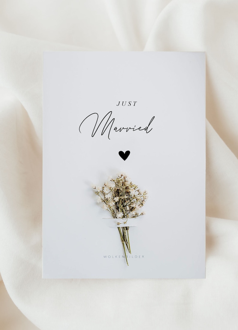 May include: A white card with the text "JUST Married" in black script font. A small black heart is below the text. A small bouquet of white flowers is below the heart. The text "WOLKENBILDER" is at the bottom of the card.