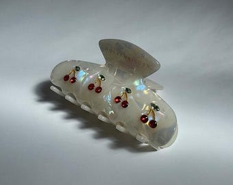 May include: Iridescent white hair claw clip adorned with red cherry and green leaf accents. The claw clip is pearlescent and features a curved design with multiple teeth for secure hair styling. The cherries are detailed with red gems.
