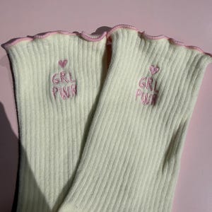 May include: A pair of cream-colored ribbed socks featuring a lettuce-edge trim in light pink. Each sock is embroidered with a small pink heart above the text GRL PWR in pink thread, positioned on the upper portion of the ribbed fabric.