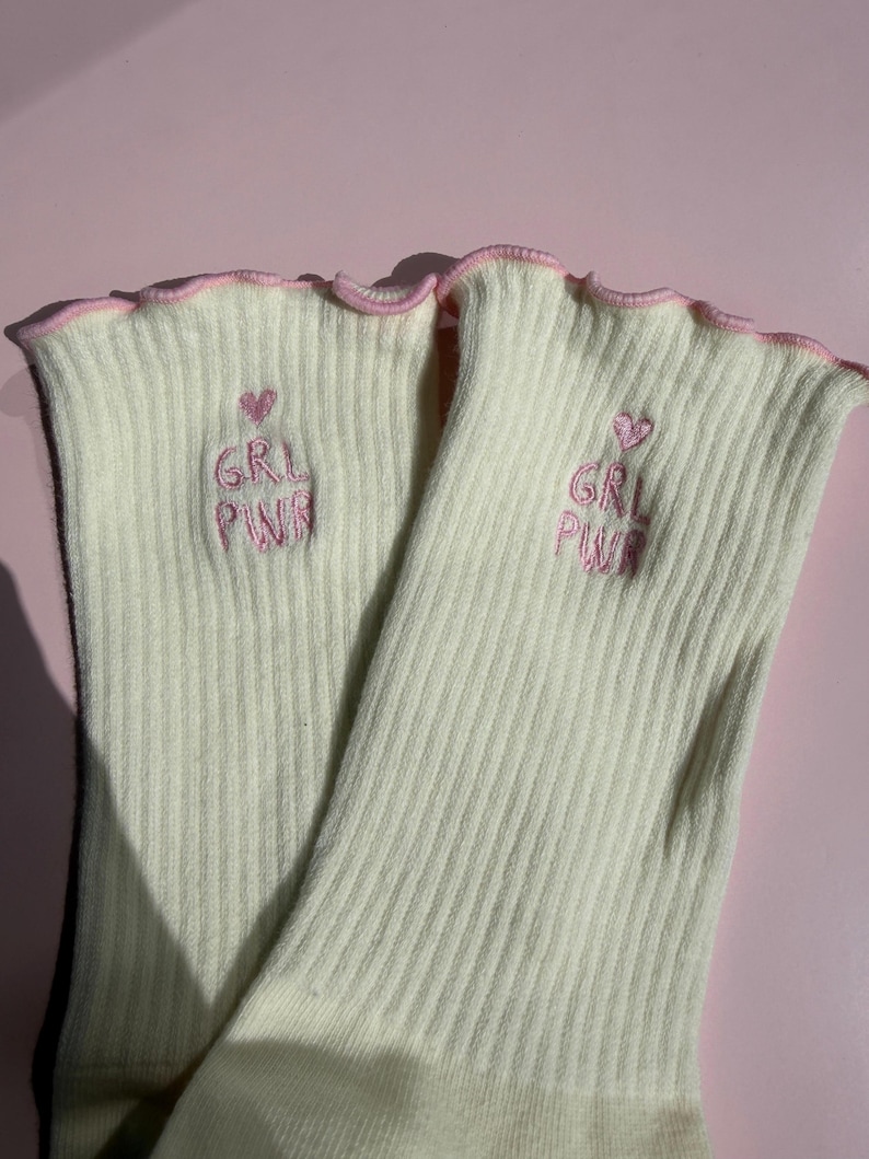 May include: A pair of cream-colored ribbed socks featuring a lettuce-edge trim in light pink. Each sock is embroidered with a small pink heart above the text GRL PWR in pink thread, positioned on the upper portion of the ribbed fabric.