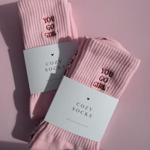 May include: A pair of light pink ribbed socks featuring the embroidered red text "YOU GO GIRL!" on the upper section. Each sock is wrapped in a white paper band printed with a small heart icon, the text "COZY SOCKS", and "WOLKENBILDER".
