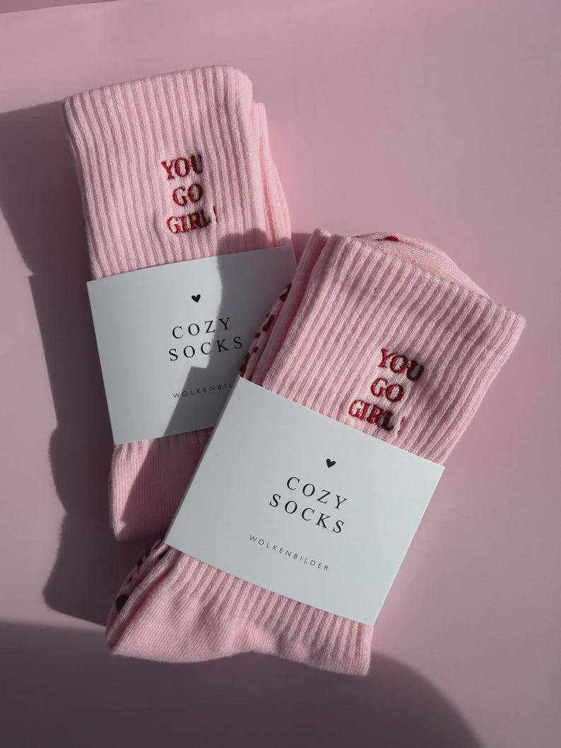 May include: A pair of light pink ribbed socks featuring the embroidered red text "YOU GO GIRL!" on the upper section. Each sock is wrapped in a white paper band printed with a small heart icon, the text "COZY SOCKS", and "WOLKENBILDER".