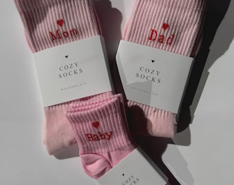 May include: Three pairs of pink ribbed knit socks. One pair has "Mom" embroidered in red with a heart, another has "Dad" embroidered in red with a heart, and the third has "Baby" embroidered in red with a heart. Each pair has a white label that reads "COZY SOCKS".
