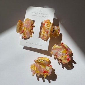 Könnte beinhalten: Vier farbenfrohe Haarspangen mit einem marmorierten Design in Rosa, Gelb und Rot. Die Spangen liegen auf einer weißen Oberfläche mit einer Visitenkarte mit der Aufschrift "WOLKENBILDER poetry and lifestyle".