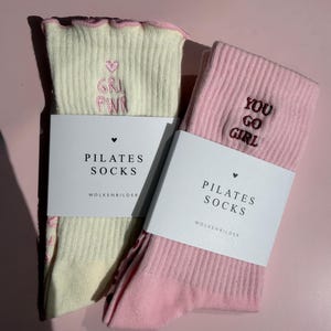 May include: Two pairs of ribbed pilates socks, one cream and one light pink, each featuring a ruffled top edge and embroidered text. The cream sock displays a pink heart and 'GRL PWR', while the pink sock features 'YOU GO GIRL'. Both are wrapped in white paper bands labeled 'PILATES SOCKS' and 'WOLKENBILDER'.