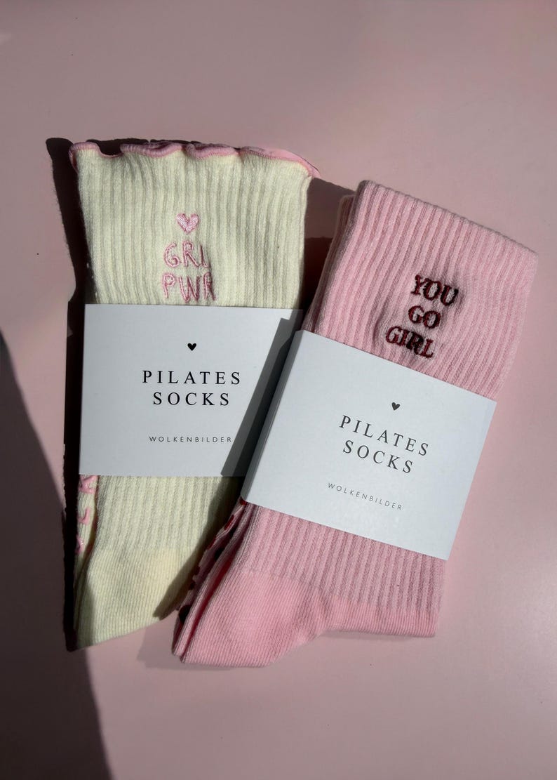 May include: Two pairs of ribbed pilates socks, one cream and one light pink, each featuring a ruffled top edge and embroidered text. The cream sock displays a pink heart and 'GRL PWR', while the pink sock features 'YOU GO GIRL'. Both are wrapped in white paper bands labeled 'PILATES SOCKS' and 'WOLKENBILDER'.