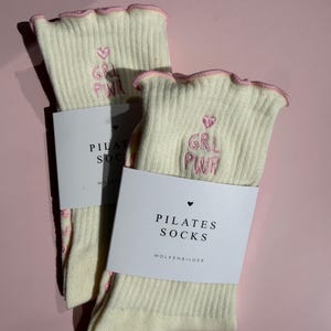 May include: A pair of cream-colored ribbed socks with pink lettuce-edge cuffs. Each sock features pink embroidered text that reads GRL PWR with a small heart above it. A white paper band is wrapped around the socks with the text PILATES SOCKS.