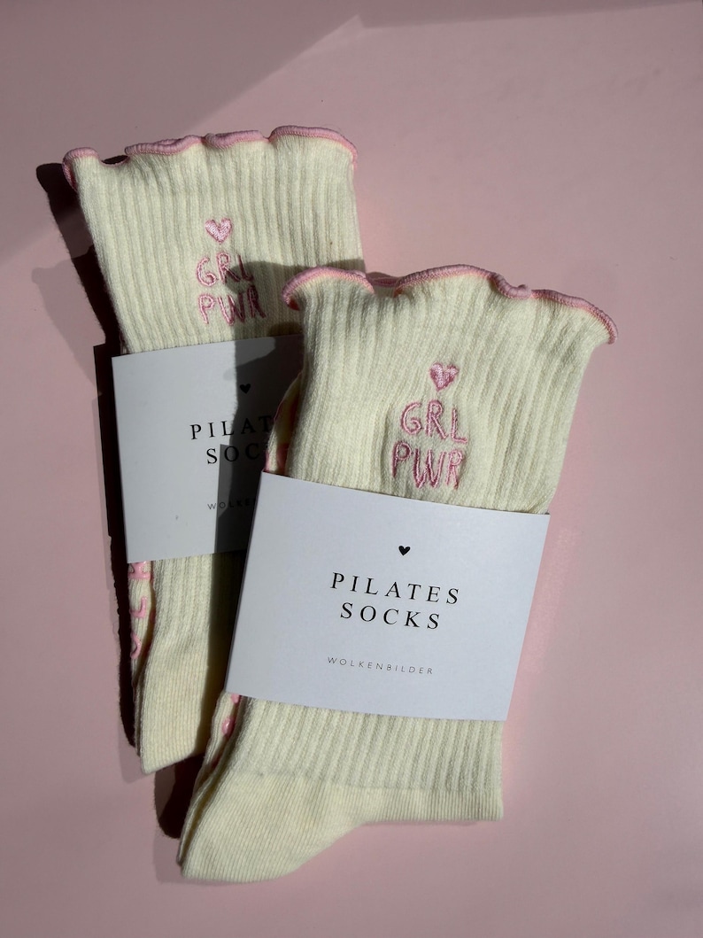 May include: A pair of cream-colored ribbed socks with pink lettuce-edge cuffs. Each sock features pink embroidered text that reads GRL PWR with a small heart above it. A white paper band is wrapped around the socks with the text PILATES SOCKS.