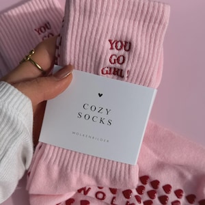 May include: A pair of light pink ribbed socks featuring the embroidered red text YOU GO GIRL! and a white paper label with the black text COZY SOCKS and WOLKENBILDER. The soles of the socks have a pattern of small red heart-shaped silicone grips.