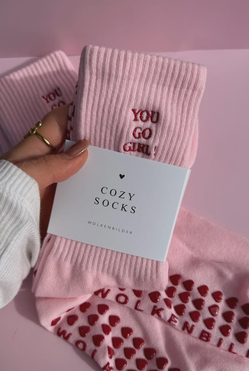 May include: A pair of light pink ribbed socks featuring the embroidered red text YOU GO GIRL! and a white paper label with the black text COZY SOCKS and WOLKENBILDER. The soles of the socks have a pattern of small red heart-shaped silicone grips.
