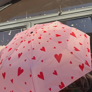 Könnte beinhalten: Ein rosa Regenschirm mit einem Muster aus roten Herzen. Der Regenschirm ist geöffnet und zeigt das Herzdesign über seine gesamte Oberfläche. Der Regenschirm hat eine hellrosa Farbe und die Herzen sind leuchtend rot.