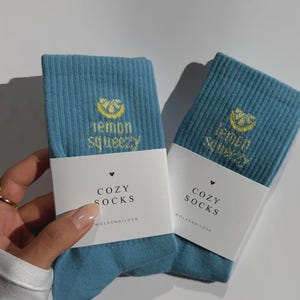 Könnte beinhalten: Zwei Paar teal-farbene Rippstrick-Socken. Jedes Paar hat ein weißes Etikett mit den Worten "COZY SOCKS" und einer Zitronengrafik mit dem Text "lemon squeezy". Der Markenname "WOLKENBILDER" ist auf dem Etikett aufgedruckt.
