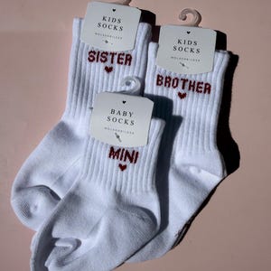 Könnte beinhalten: Drei Paar weiße Rippsocken mit rotem Sticktext und Herzsymbolen. Zwei Paare sind mit Kids Socks und den Wörtern Sister und Brother beschriftet, während das kleinere Paar mit Baby Socks und dem Wort Mini versehen ist. Jedes Paar hat einen Aufhänger.