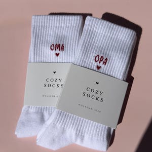 Könnte beinhalten: Ein Paar weiße Rippstricksocken mit der roten Stickerei OMA und OPA sowie kleinen Herzsymbolen. Jede Socke ist mit einer hellgrauen Papierbanderole versehen, auf der der Text COZY SOCKS und der Markenname WOLKENBILDER in Schwarz gedruckt sind.