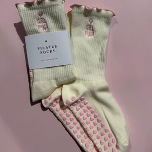 May include: A pair of cream-colored ribbed pilates socks with pink lettuce-edge cuffs. The socks feature pink embroidered text reading GRL PWR with a heart above it. The soles have pink heart-shaped silicone grips. A white paper label reads PILATES SOCKS.