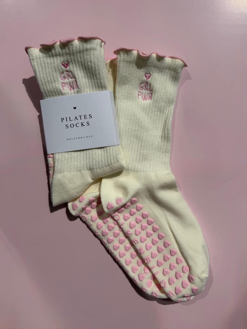 May include: A pair of cream-colored ribbed pilates socks with pink lettuce-edge cuffs. The socks feature pink embroidered text reading GRL PWR with a heart above it. The soles have pink heart-shaped silicone grips. A white paper label reads PILATES SOCKS.