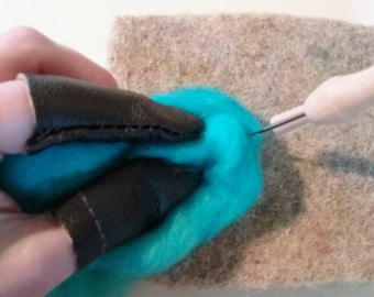 May include: A close-up view of a craft project in progress. A vibrant turquoise wool ball is being shaped with a needle tool. The crafter wears black leather finger protectors. The project rests on a gray felt surface.