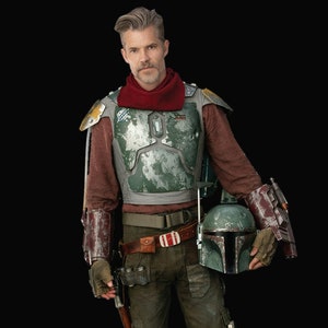 May include: A person wearing a brown, red and green costume with a helmet in hand. The costume has a weathered, worn look.