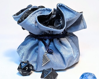 May include: A light blue velvet dice bag with a black and white geometric pattern. The bag is tied with a dark blue cord and has a silver charm. There are five dice on a white surface, including a black and silver d20, a blue and silver d20, a clear d6 with blue and gold accents, a black and silver d12, and a turquoise d4.