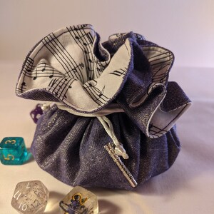 May include: Purple fabric dice bag with a white musical note lining. The bag has a silver drawstring and a silver charm. Four dice are scattered around the bag, two are clear with silver glitter, one is blue with gold numbers, and one is teal with gold numbers.