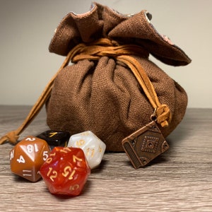 May include: A brown fabric dice bag with a leather drawstring and a metal book charm. The bag is filled with dice, including a red d20, a white d20, a brown d20, and a black d6.