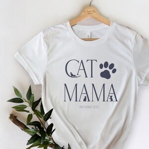May include: White t-shirt with a navy blue graphic that says "Cat Mama" with a paw print and cat silhouettes. The text "Proverbs 12:10" is also included on the shirt.