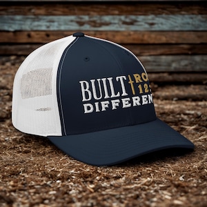 May include: Navy blue trucker hat with a white mesh back and a curved brim. The front of the hat features the text "BUILT DIFFERENT" in white, with "ROM 12:2" in gold, and a gold cross.