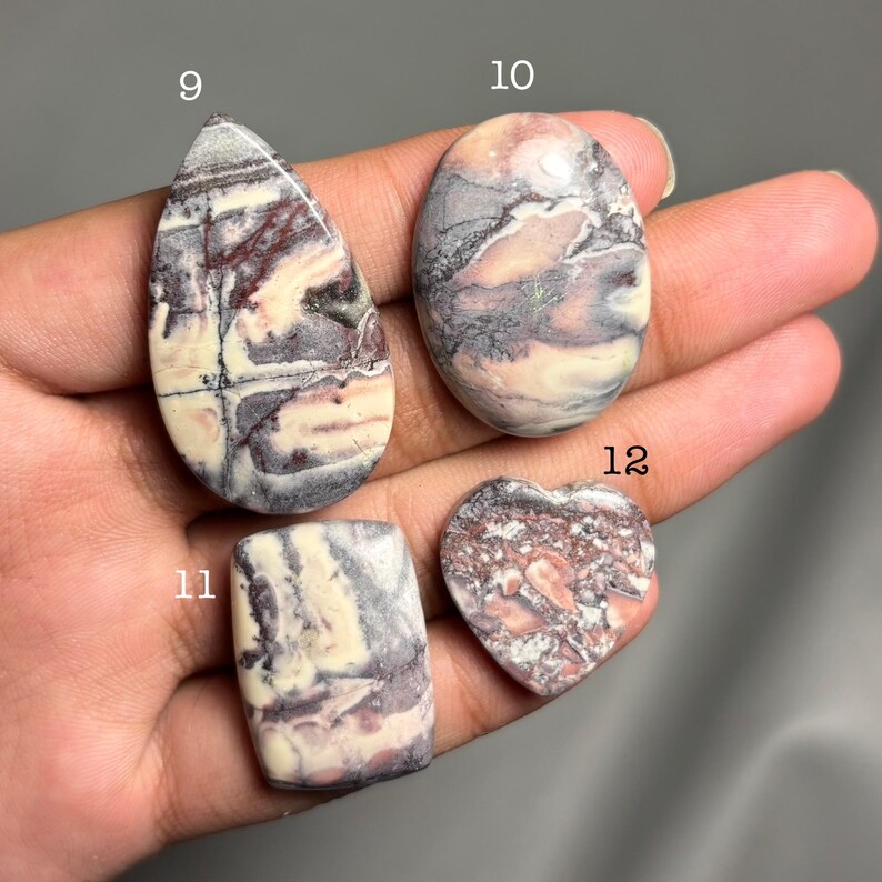 May include: Four polished gemstones in various shapes: a teardrop, an oval, a rectangle, and a heart. The stones display a mix of colors, including cream, purple, and reddish-brown, with unique veining patterns. Each stone is numbered.