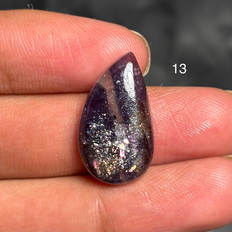 May include: A teardrop-shaped gemstone with a deep purple hue, featuring shimmering inclusions of silver, gold, and iridescent colors. The number "13" is visible in white. The stone is held between fingers.