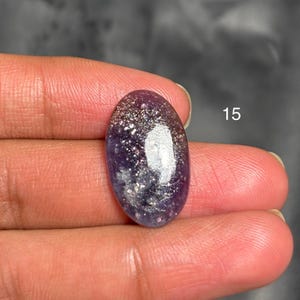 May include: An oval-shaped gemstone with a deep purple base, speckled with silver and white inclusions. The stone has a polished, glossy surface. The number "15" is visible in the background.