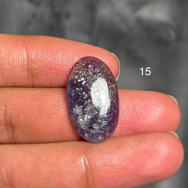 May include: An oval-shaped gemstone with a deep purple base, speckled with silver and white inclusions. The stone has a polished, glossy surface. The number "15" is visible in the background.
