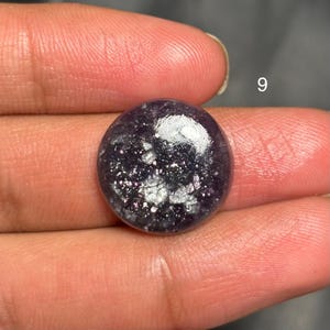 May include: A round, polished gemstone with a deep purple hue and shimmering silver inclusions. The stone is held between fingers, with the number "9" visible in the background. The gem has a smooth, reflective surface.