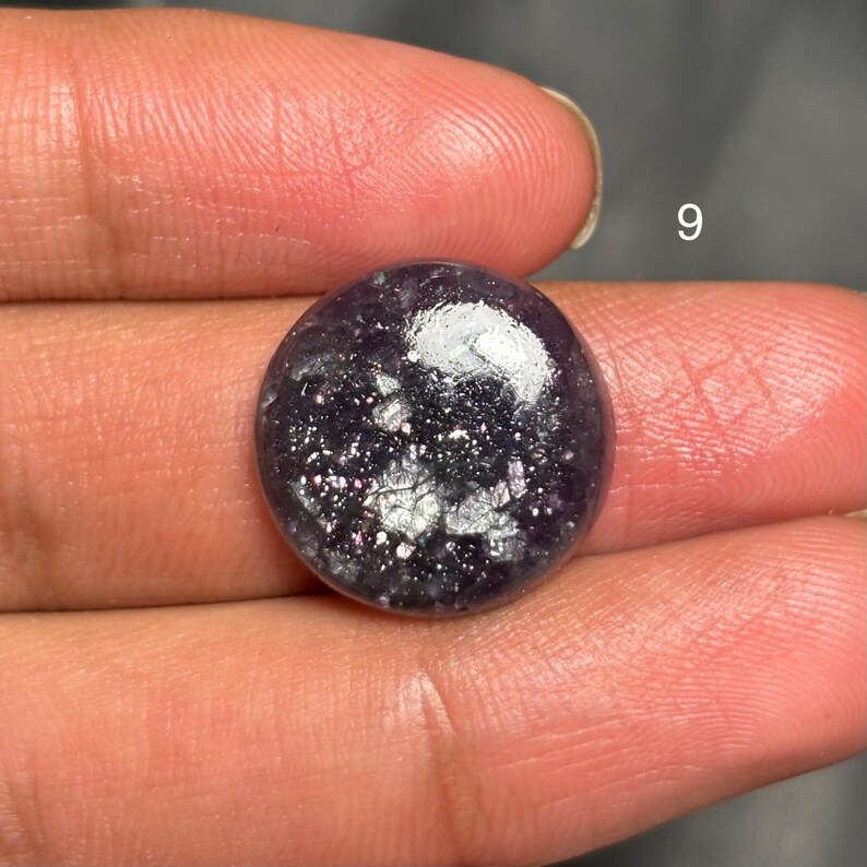 May include: A round, polished gemstone with a deep purple hue and shimmering silver inclusions. The stone is held between fingers, with the number "9" visible in the background. The gem has a smooth, reflective surface.