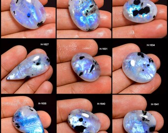 May include: Nine polished moonstone cabochons with iridescent blue and white flashes and black inclusions. Each stone is a different shape, including oval and teardrop. The stones are held in a hand.