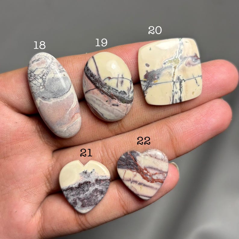 May include: Five polished gemstones in various shapes: oval, square, and heart. The stones feature a cream-colored base with intricate patterns in gray, brown, and pink. Each stone is numbered for identification.