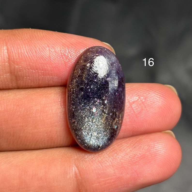 May include: An oval-shaped gemstone with a polished surface. The stone features a blend of deep purple and silver hues, with sparkling inclusions. The number "16" is visible in white. The gemstone is held in a hand.