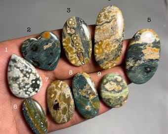 May include: A collection of nine polished gemstones in various shapes, including teardrop, heart, and oval. The stones display a mix of colors, such as green, brown, and cream, with unique patterns and textures. The gemstones are held in a hand.