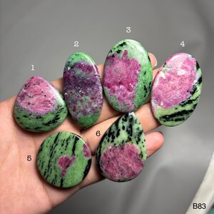 Può includere: Una collezione di sei pietre preziose di rubino zoisite lucide in varie forme, tra cui ovali, a goccia e rotonde. Le pietre presentano un vivace mix di colori rosa, verde e nero. Le pietre preziose sono tenute in una mano.