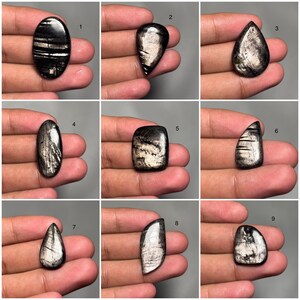 May include: A collection of nine polished black gemstones with iridescent silver inclusions. Each stone is a different shape, including oval, teardrop, and rectangular. The stones are held in a hand against a gray background, showcasing their unique patterns and textures.