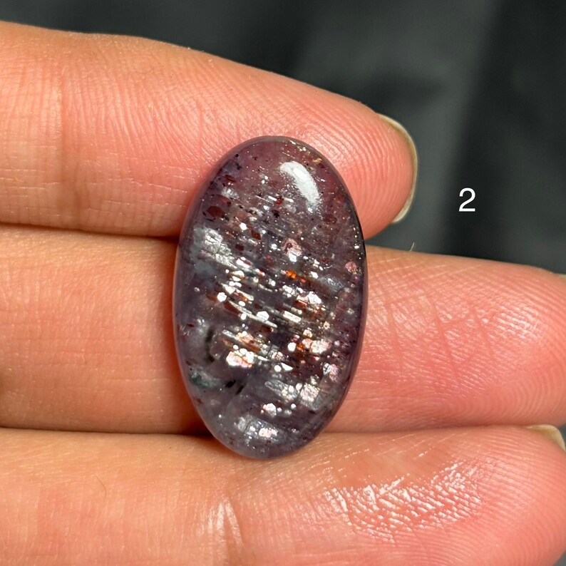 May include: An oval-shaped gemstone with a dark purple base color and shimmering silver and copper inclusions. The polished surface reflects light, creating a sparkling effect. The gemstone is held between fingers.