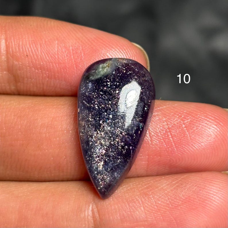 May include: A teardrop-shaped, polished gemstone with a deep purple hue, speckled with shimmering silver and gold inclusions. The number "10" is visible in white. The stone is held between fingers.
