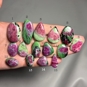 May include: Fifteen ruby zoisite gemstones in diverse shapes, such as oval and teardrop. The stones exhibit a mix of vibrant pink, deep red, and green colours, with some featuring black inclusions. The gemstones are displayed on a neutral background.