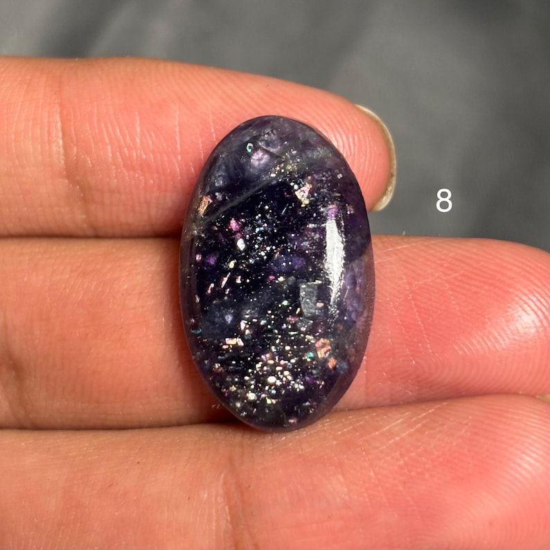 May include: An oval-shaped, polished gemstone with a deep purple hue, speckled with iridescent flecks. The stone is held in a hand, with the number "8" visible in the background.