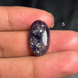 May include: An oval-shaped, polished gemstone with a deep purple hue, speckled with iridescent flecks of silver and hints of pink. The stone is smooth and reflective, showcasing a natural, unique pattern. The number "5" is visible in the background.