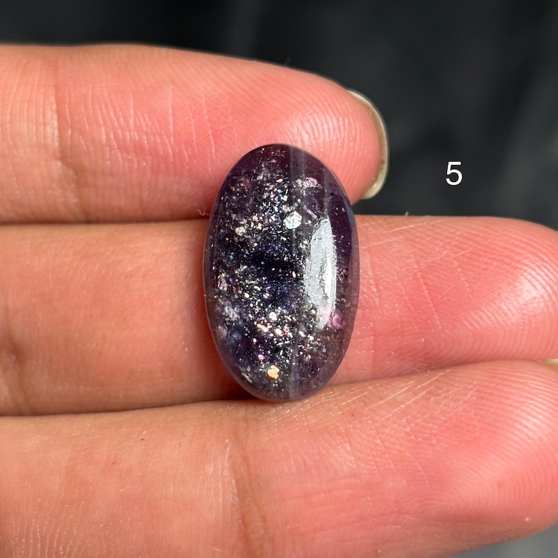 May include: An oval-shaped, polished gemstone with a deep purple hue, speckled with iridescent flecks of silver and hints of pink. The stone is smooth and reflective, showcasing a natural, unique pattern. The number "5" is visible in the background.