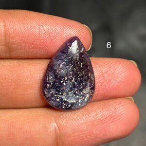 May include: A teardrop-shaped, polished gemstone with a deep purple hue, speckled with silver and iridescent glitter. The number "6" is visible in the background. The stone is held between fingers.