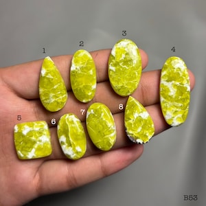 Puede incluir: Una colección de ocho piedras preciosas pulidas de varias formas, incluyendo ovaladas, en forma de lágrima y cuadradas. Las piedras son de un color verde-amarillo vibrante con vetas blancas. Las piedras preciosas se muestran en una mano.